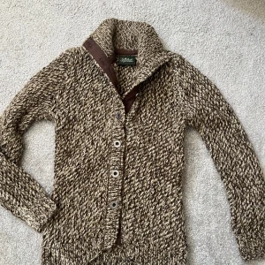 Stickad kofta från Peak Performance - Säljer en chunky stickad kofta från Peak Performance i beige och bruna toner. Koftan har grovstickat mönster, bred krage med bruna detaljer och metallknappar framtill. Perfekt för kyliga dagar och riktigt snygg till jeans eller chinos.