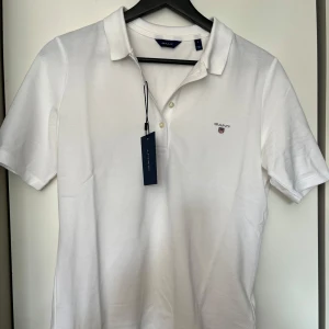 Vit pikétröja från GANT, storlek S - Fräsch vit pikétröja från GANT i klassisk design med krage och två knappar framtill. Tröjan har korta ärmar och GANT-logga broderad på bröstet. Tillverkad i mjuk bomull som andas, perfekt för vår och sommar. Snygg och enkel att matcha med jeans eller shorts.