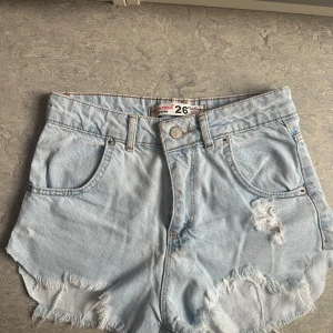 Ljusblå jeansshorts från Noa&Noa - Snygga ljusblå jeansshorts från Noa&Noa med vintage vibe. Den har fransiga kanter och slitningar framtill för en cool look. Klassiska fem fickor och knappstängning. Perfekta för varma dagar och chill stil.