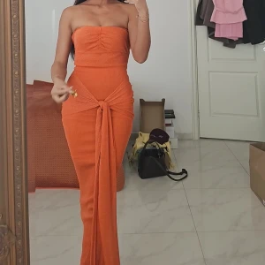 Orange långklänning med knyt - Säljer en snygg orange axelbandslös långklänning med glittrigt tyg och knyt framtill i midjan. Klänningen har en slim passform och en cool omlott-detalj som ger extra edge, tillkommer även sjal med. Perfekt för dig som vill sticka ut med färg och stil på nästa event.