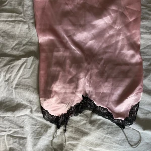 Rosa satin linne med spetsdetaljer - Supersnygg kort klänning i rosa satin med svarta spetsdetaljer och tunna axelband. Baksidan har genomskinlig svart spets med dekorativa knappar och blommigt mönster. Perfekt för dig som gillar romantisk och glamorös stil.