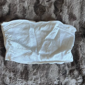 Vit bandeau tubtopp med smock - Säljer en vit bandeau tubtopp med smockad baksida och små volangdetaljer upptill och nedtill. Toppen är axelbandslös och har en mjuk, lätt struktur som gör den skön att bära. 