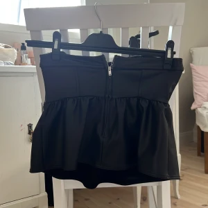 Svart volang korsett-topp med dragkedja - Säljer en svart korsett-topp med snygg volang nedtill och dragkedja framtill. Toppen är axelbandslös och har en cool, strukturerad look som ger extra edge till outfiten. Materialet är slätt och har en lätt glansig finish som känns riktigt trendig.