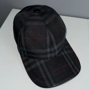 Rutig svart keps från Burberry - Snygg keps från Burberry i svart med diskret rutig mönster i grått och röda linjer. Klassisk form med böjd skärm och justerbar rem bak. Tillverkad i polyester med insida i mjukt tyg. Perfekt för dig som vill ha en stilren accessoar med lyxig känsla.