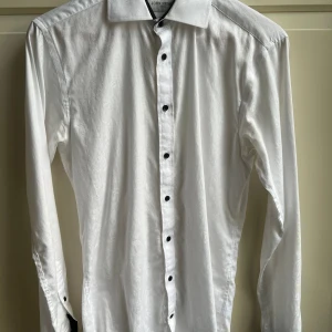 Vit skjorta med svarta detaljer John Henric - Snygg vit skjorta från John Henric med subtilt mönster i tyget och svarta knappar. Manschetterna har coola svarta insidor som ger en unik touch. Skjortan har klassisk krage och är långärmad, perfekt för dig som gillar stilrena plagg med lite edge.