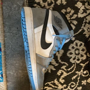 Nike Air Jordan 1 High blå/grå - Nike Air Jordan 1 High sneakers i en clean mix av ljusgrå mocka och vitt skinn, med svarta swoosh-detaljer och blå snören. Yttersulan är blå och skon har den klassiska Air Jordan-loggan på sidan. Perfekt för dig som vill sticka ut med en ikonisk siluett.