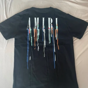 Svart AMIRI t-shirt med färgstänk - Svart t-shirt från AMIRI med coolt tryck av loggan och färgstänk i blått, orange, grönt och vitt både fram och bak. Klassisk rund hals och korta ärmar. Materialet är mjuk bomull som känns skönt mot huden. Perfekt för dig som gillar streetwear och vill sticka ut.