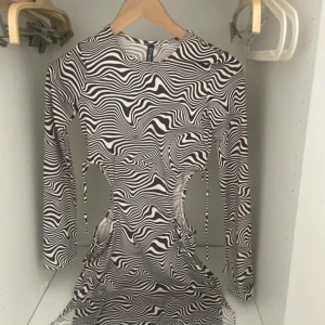 Zebramönstrad cut-out klänning - Säljer en svartvit zebramönstrad kortklänning från H&M Divided i storlek XS. Klänningen har långa ärmar, coola cut-outs i midjan med snörningsdetaljer och är gjord i ett stretchigt syntetmaterial. Perfekt för dig som gillar statement-plagg och edgy stil.