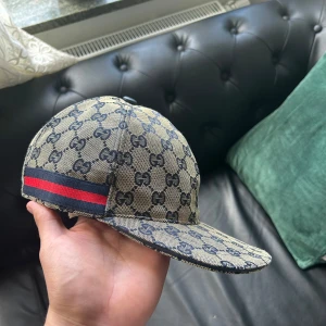 Gucci keps med GG-mönster och röd-blå rand - Säljer en beige keps från Gucci med ikoniskt GG-mönster i mörkblått och en snygg röd-blå rand på sidan. Kepsen är tillverkad i bomull och har en klassisk böjd skärm. Insidan är ljus och har Gucci-tag. Perfekt för dig som gillar lyxiga accessoarer.