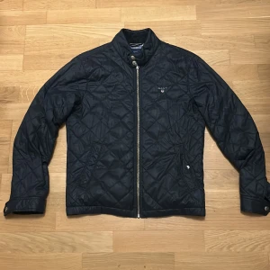 Svart quiltad jacka från GANT - Quiltad svart jacka från GANT med snyggt rutigt foder och ståkrage i manchester. Jackan har dragkedja framtill, tryckknappar vid ärmslut och två fickor. Perfekt för dig som gillar klassisk stil med en modern twist. Vänstra fickan har en trasig knapp 