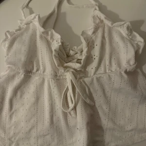 Vit linnetopp med snörning och volang - Supersöt vit linnetopp med smala axelband, volangdetaljer vid halsen och snygg snörning framtill. Toppen har ett broderat hålmönster som ger en chill och romantisk vibe.