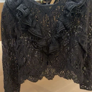 Svart spetsblus med volanger - Supersnygg svart blus i spets med unika broderade detaljer och volanger framtill. Långärmad och med en cool, genombruten look som ger extra edge till outfiten. Perfekt för dig som vill sticka ut med stilrena detaljer och mönster.