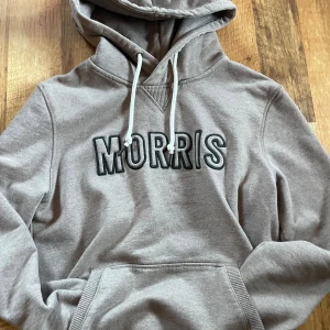 Beige hoodie från Morris med tryck - Snygg beige hoodie från Morris med broderat logotyp-tryck i mörkgrönt på bröstet. Tröjan har huva med vita snören och en klassisk känguruficka framtill. Mjuk och skön bomullsmix, perfekt för chill dagar. Passar dig som gillar stilren streetwear.