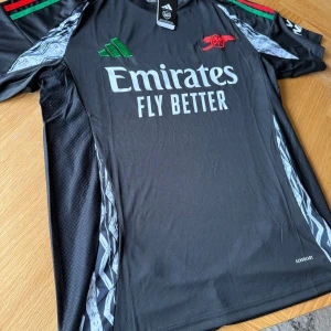 Arsenal från Adidas - Säljer en svart fotbollströja från Adidas med gröna och röda detaljer. Tröjan har korta ärmar och ett tryck med texten 'Emirates Fly Better' på framsidan. Perfekt för fotbollsfans som vill ha en stilren look på planen.