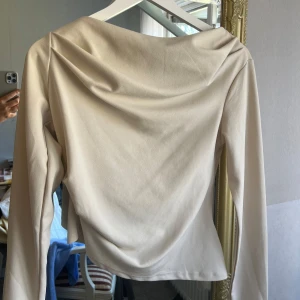 Beige långärmad topp - Säljer en elegant beige topp med långa ärmar och draperad halsringning. Perfekt för en stilren look. Passar bra till både jeans och kjol.