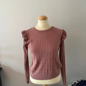 Rosa stickad tröja med volangdetaljer - Säljer en söt rosa stickad tröja med volangdetaljer på axlarna. Tröjan har ett kabelstickat mönster och långa ärmar. Perfekt för kyligare dagar!