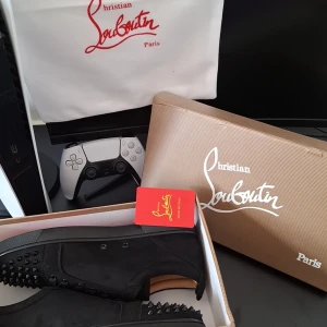Svarta sneakers med nitar från Christian Louboutin - Snygga svarta sneakers från Christian Louboutin med ikonisk röd sula och nitar på ovansidan. Skorna har snörning och är tillverkade i mocka, vilket ger en lyxig känsla. Perfekta för att ge din outfit en edgy touch.