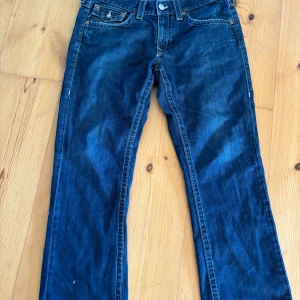 Mörkblå jeans från True Religion - Säljer ett par mörkblå jeans från True Religion med klassisk femficksdesign och snygga sömmar på bakfickorna. Jeansen har en knappgylf och är tillverkade i USA. Perfekta för en avslappnad stil.