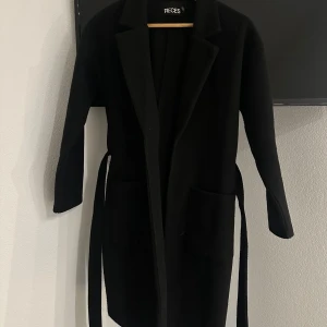 Svart kappa från Pieces - Snygg svart kappa från Pieces med långärmad design och bälte i midjan. Perfekt för höst och vår, med en klassisk look som passar till många tillfällen. Kappan har en stilren och enkel design med fickor framtill.