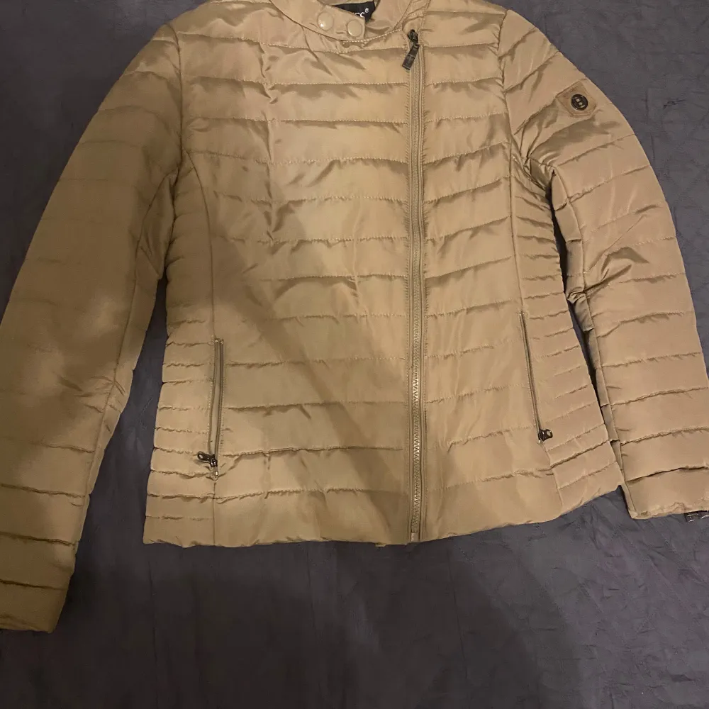 Säljer en snygg beige quiltad jacka från Hollies. Jackan har en dragkedja framtill och långa ärmar. Perfekt för kyligare dagar med sin stilrena design och bekväma passform.. Takit.