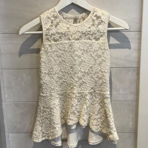 Vit spetsblus - Säljer en elegant vit spetsblus med ärmlös design och vacker spetsdetalj. Blusen har en lätt utsvängd nederdel och en knapp i nacken för enkel påklädning. Perfekt för en stilren look.  Nästan aldrig använd.