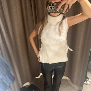 Vit ribbad ärmlös topp - Säljer en stilren vit ribbad topp med hög krage och knytband i sidorna. Perfekt för lager-på-lager eller som den är. Passar till både jeans och kjol för en chic look.