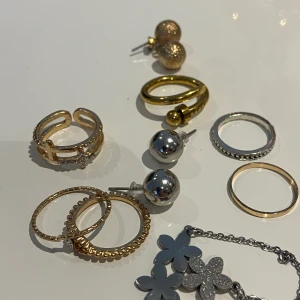 Smyckeset med örhängen och ringar - Ett elegant smyckeset med olika örhängen och ringar. Setet innehåller guld- och silverfärgade ringar samt örhängen i både guld och silver. Några av örhängena har glittriga detaljer och det finns även ett halsband med blommotiv.