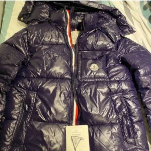 Mörkblå pufferjacka från Moncler - Säljer en snygg mörkblå pufferjacka från Moncler med en glansig finish. Jackan har en dragkedja i kontrastfärg och en praktisk bröstficka med Moncler-logga. Perfekt för kyliga dagar!