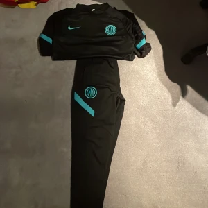 Nike dri fit - Säljer ett svart träningsset från Nike med Inter-logga. Tröjan har en halv dragkedja och turkosa detaljer på ärmarna. Byxorna har en matchande turkos detalj och logga. Perfekt för fotbollsträning eller annan sportaktivitet.
