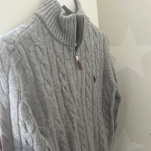 Grå kabelstickad tröja från Polo Ralph Lauren - Säljer en stilren grå kabelstickad tröja från Polo Ralph Lauren. Tröjan har en dragkedja framtill och en hög krage för extra värme. Perfekt för kyligare dagar med sin klassiska design och mjuka material.
