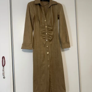 Beige långklänning från Zara - Snygg beige långklänning från Zara med knappar hela vägen ner och en stilren krage. Klänningen har långa ärmar och en rynkad detalj framtill som ger en fin siluett. Perfekt för en chic och elegant look.