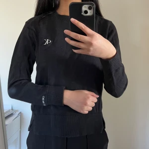 Svart tröja från Calvin Klein - Snygg svart finstickad tröja från Calvin Klein med diskret logga på bröstet. Tröjan har långa ärmar och ribbade muddar vid ärmslut och nederkant. Perfekt för en stilren look.