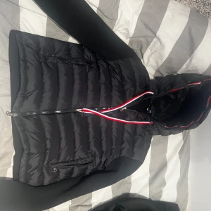 Svart dunjacka från Moncler - Snygg svart dunjacka från Moncler med huva och dragkedja. Jackan har ribbade ärmar och en stilren design med Moncler-logga på ärmen. Perfekt för kyligare dagar. Liten i storlek samt oanvänd 