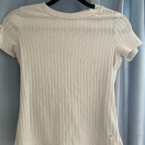 Vit ribbad topp från Lindex - Säljer en stilren vit ribbad topp från Lindex. Den har korta ärmar och en klassisk rund halsringning. Perfekt för en enkel och snygg look. Den är egentligen för barn men passar xxs och xs också 