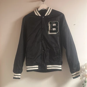 Svart bomberjacka - Snygg svart bomberjacka med vita ribbade muddar och krage. Jackan har en broderad bokstav på bröstet och stängs med knappar. Jackan har två fickor på framsidan. Perfekt för en cool och avslappnad stil. Storlek XS men funkar för S och M också med skulle jag säga. Den ger lite Lana Del Rey vibes 💞
