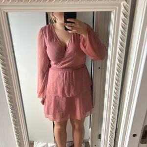 Rosa omlottklänning - Säljer en söt rosa omlottklänning med v-ringning och långa ärmar. Klänningen har en volangdetalj i nederkant och är perfekt för vår och sommar. Den har en smickrande passform och är gjord i ett lätt och luftigt material.