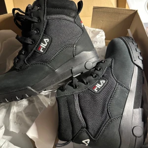 Svarta kängor från Fila - Snygga svarta kängor från Fila med robust sula och snörning. Perfekta för en cool och bekväm stil. Märkeslogga på sidan och sulan ger en extra touch. Nya i lådan