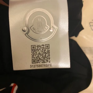 Svart pikétröja från Moncler - Säljer en stilren svart pikétröja från Moncler med klassiska röd-vita detaljer på kragen och ärmsluten. Tröjan har en broderad Moncler-logga på bröstet. Perfekt för en sportig och elegant look.