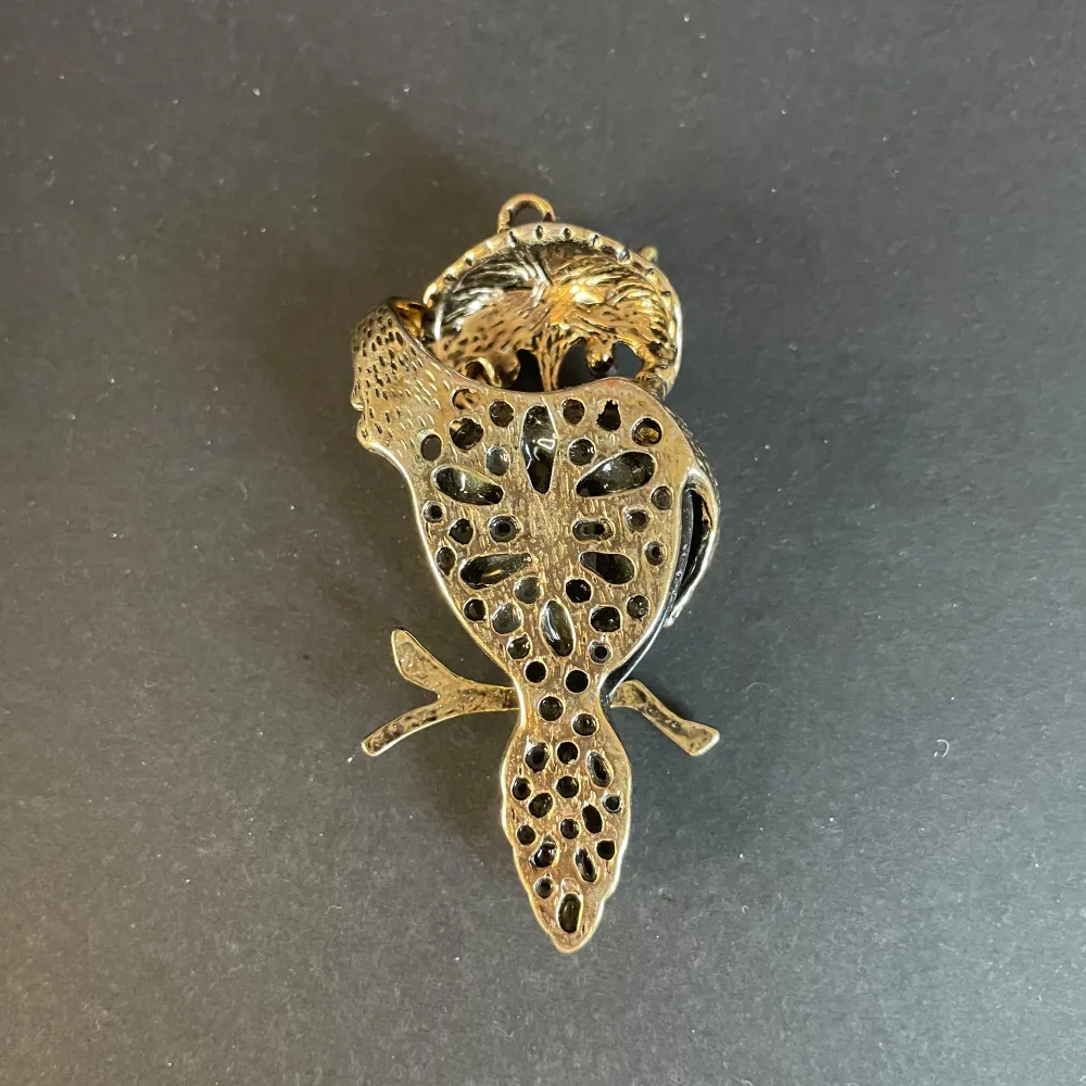 Unikt halsband med en uggla i guld och svart. Ugglan har ett detaljerat mönster och hänger från en gren. Kedja till smycket saknas . Asusteet.
