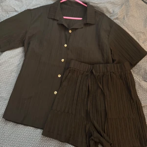 Svart kortärmad skjorta med shorts - Snygg svart kortärmad skjorta med tillhörande shorts. Skjortan har en klassisk krage och är tillverkad i ett lätt material som ger en bekväm passform. Perfekt för en stilren look. Använd 1 gång. 