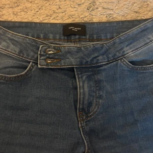 Blå jeansbyxor från Vero Moda - Snygga blå jeansbyxor från Vero Moda med låg midja och bootcut-stil. Byxorna har en klassisk femficksdesign och stängs med två knappar framtill. Perfekta för en avslappnad look. Aldrig använda