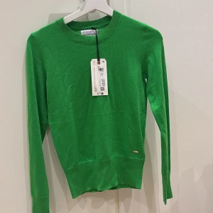 Grön tröja från Holly & Whyte - Snygg grön tröja från Holly & Whyte med långa ärmar och dekorativa knappar vid ärmsluten. Perfekt för en stilren look. Tröjan har en rund halsringning och är tillverkad i ett mjukt material.