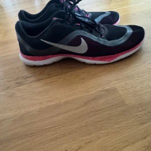 Svarta och rosa Nike sneakers - Snygga Nike sneakers i svart och rosa med grå detaljer. Skorna har en vit sula och klassisk snörning. Perfekta för träning eller en aktiv vardag.