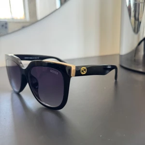 Svarta solglasögon från Gucci - Snygga svarta solglasögon från Gucci med guldiga detaljer på bågarna. De har en klassisk fyrkantig form och Gucci-loggan på sidorna. Kopia, helt nya. Fodral ingår inte! 