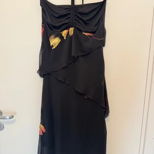 Svart axelbandslös klänning med blommönster - Elegant svart axelbandslös klänning med vackra blommönster i rött och gult. Klänningen har volangdetaljer och en smickrande omlott-design. Perfekt för en stilfull kväll.