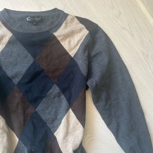 Grå argyle-mönstrad tröja från Coollike - Säljer en stilren grå tröja från Coollike med klassiskt argyle-mönster i beige, brun och mörkblå. Tröjan har långa ärmar och rund halsringning. 