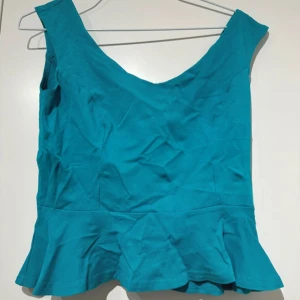 Turkos ärmlös topp - Snygg turkos ärmlös topp med volangdetalj i nederkant. Perfekt för en somrig look. Toppen har en v-ringad design och är tillverkad i ett lätt och bekvämt material.