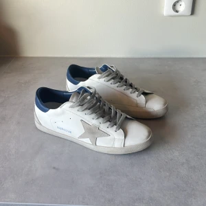 Golden Goose - Snygga vita sneakers från Golden Goose. Skorna har grå snörning och en mörkblå häl. Perfekta för en stilren och avslappnad look. Inget kvitto, påse tillkommer.