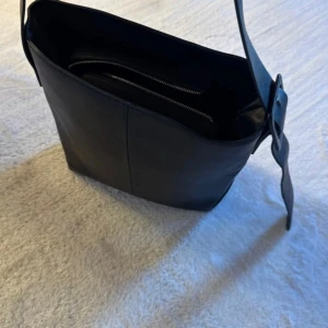 Svart axelväska i imitation av skinn med spänne - Snygg svart axelväska  med bred axelrem och stilrent spänne. Väskan har ett rymligt huvudfack och innerfack med dragkedja, perfekt för att hålla ordning på dina grejer. Enkel och clean design som funkar till många olika outfits. 35x30 och nypris är 499