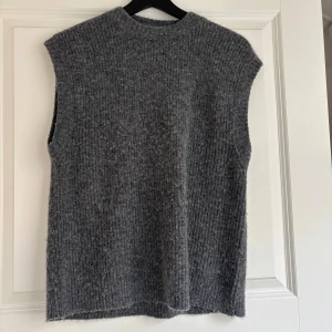 Mörkgrå stickad väst från Norr - Mysig mörkgrå stickad väst med ribbad struktur och hög hals. Så fin att ha över en skjorta eller t shirt. Västen är rak i modellen. Stl onesize.